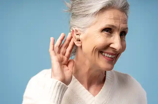 Ältere Frau mit Hörgerät lächelt und hält Hand ans Ohr.