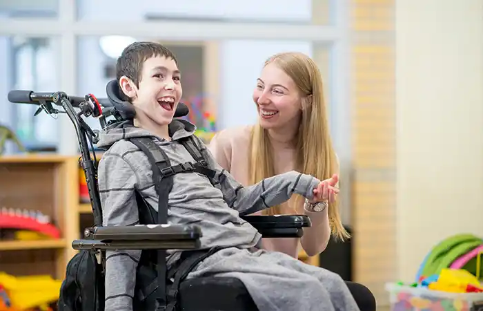 Junge im Rollstuhl lacht mit Betreuerin in einem Spielzimmer.