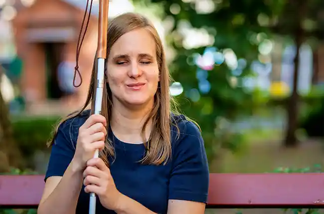 Blinde Frau mit Langstock sitzt lächelnd auf einer Parkbank.
