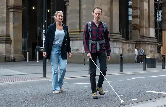 Blinder Mann mit Langstock überquert Straße, Frau begleitet ihn.