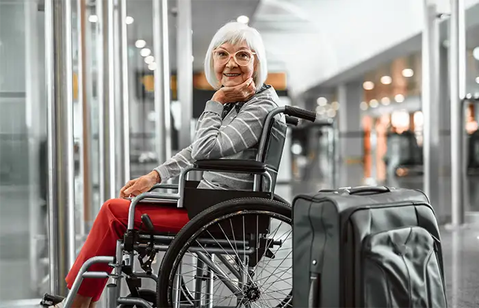 Ältere Frau im Rollstuhl sitzt lächelnd neben ihrem Koffer am Flughafen.