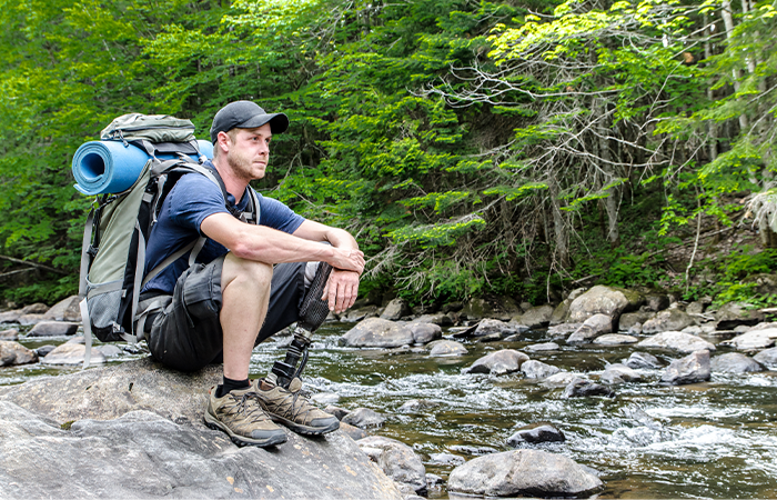 Wanderer mit Trekkingrucksack und Prothese ruht an einem felsigen Bachufer im Grünen.