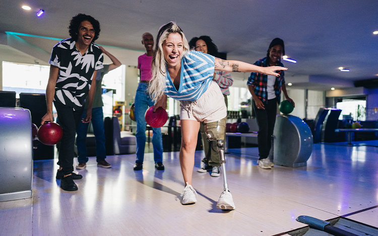 Frau mit Beinprothese wirft lachend eine Bowlingkugel, Freunde stehen hinter ihr